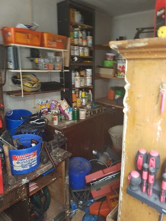 Unaufgeräumte Werkstatt mit Werkzeug, Farbeimern, Regalen und verschiedenen Utensilien.